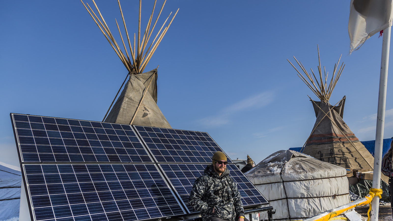 A man sits on a solar panel next to Indigenous teepees.