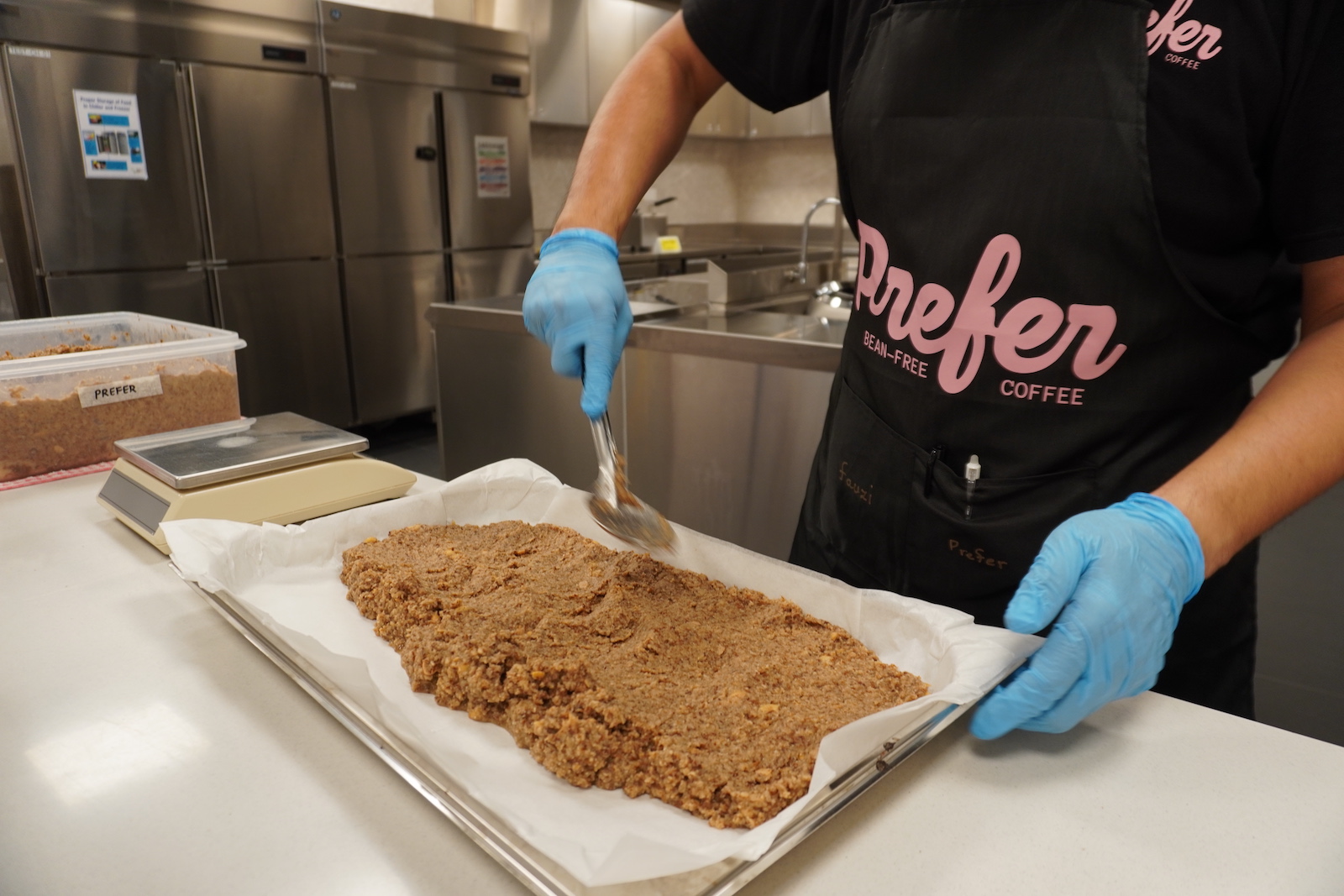 A person in a black apron with the word "Prefer" on it smooths out brown grounds in a baking hseet