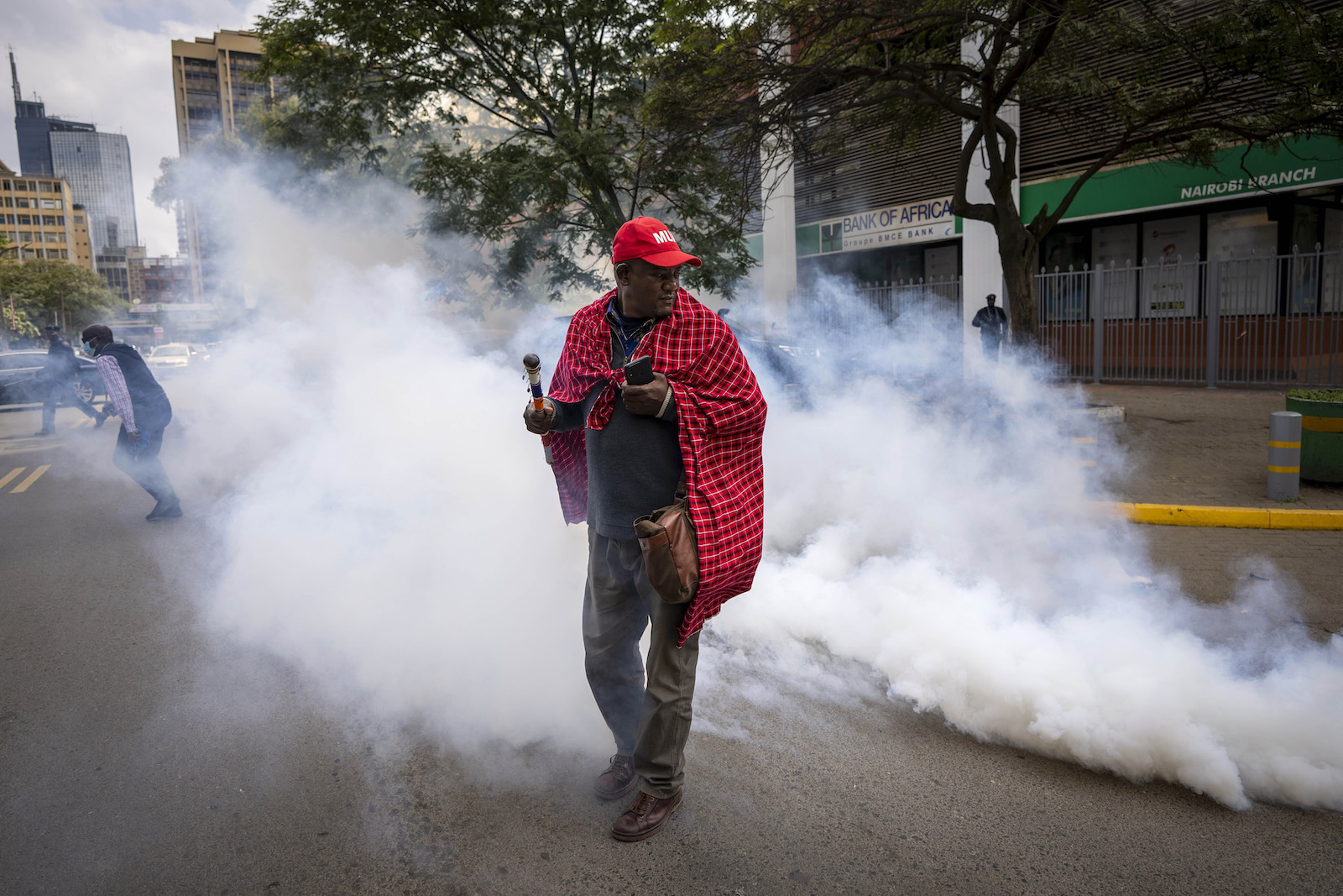 A person in a red baseball cap and draped in a red traditional Maasai cloth walks in the middle of a city street as white clouds of gas billow around him
