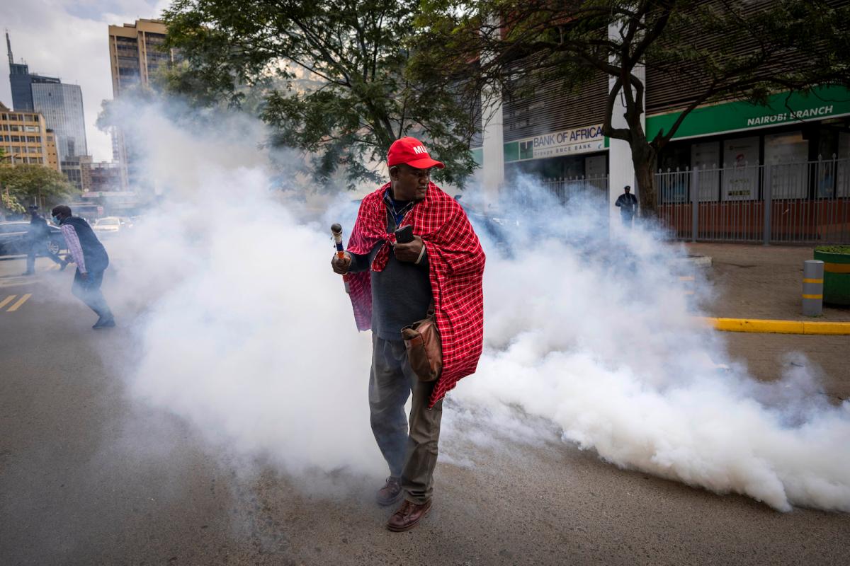 A person in a red baseball cap and draped in a red traditional Maasai cloth walks in the middle of a city street as white clouds of gas billow around him