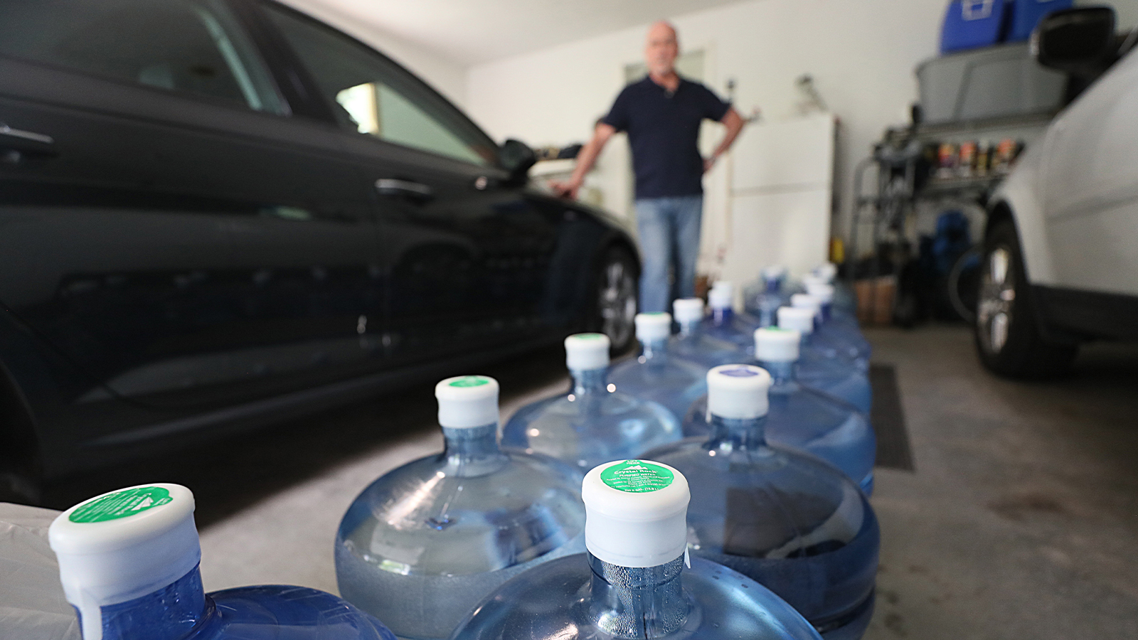 A man stands in his garage behind a long row of water bottles delivered to his home because the local water supply is contiaminated with PFAS.