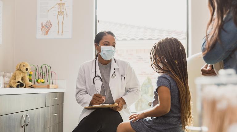 A small child is seen from behind, talking to a female doctor wearing a face mask. Her mother stands nearby.