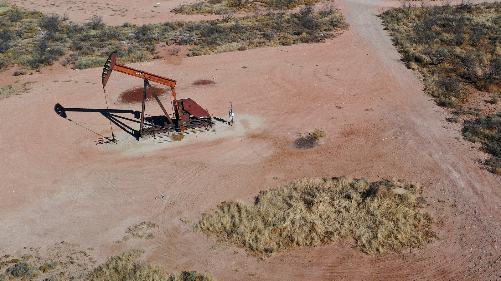An aerial view of reddish sand and a red oil rig in the middle of two areas of scrub brush.