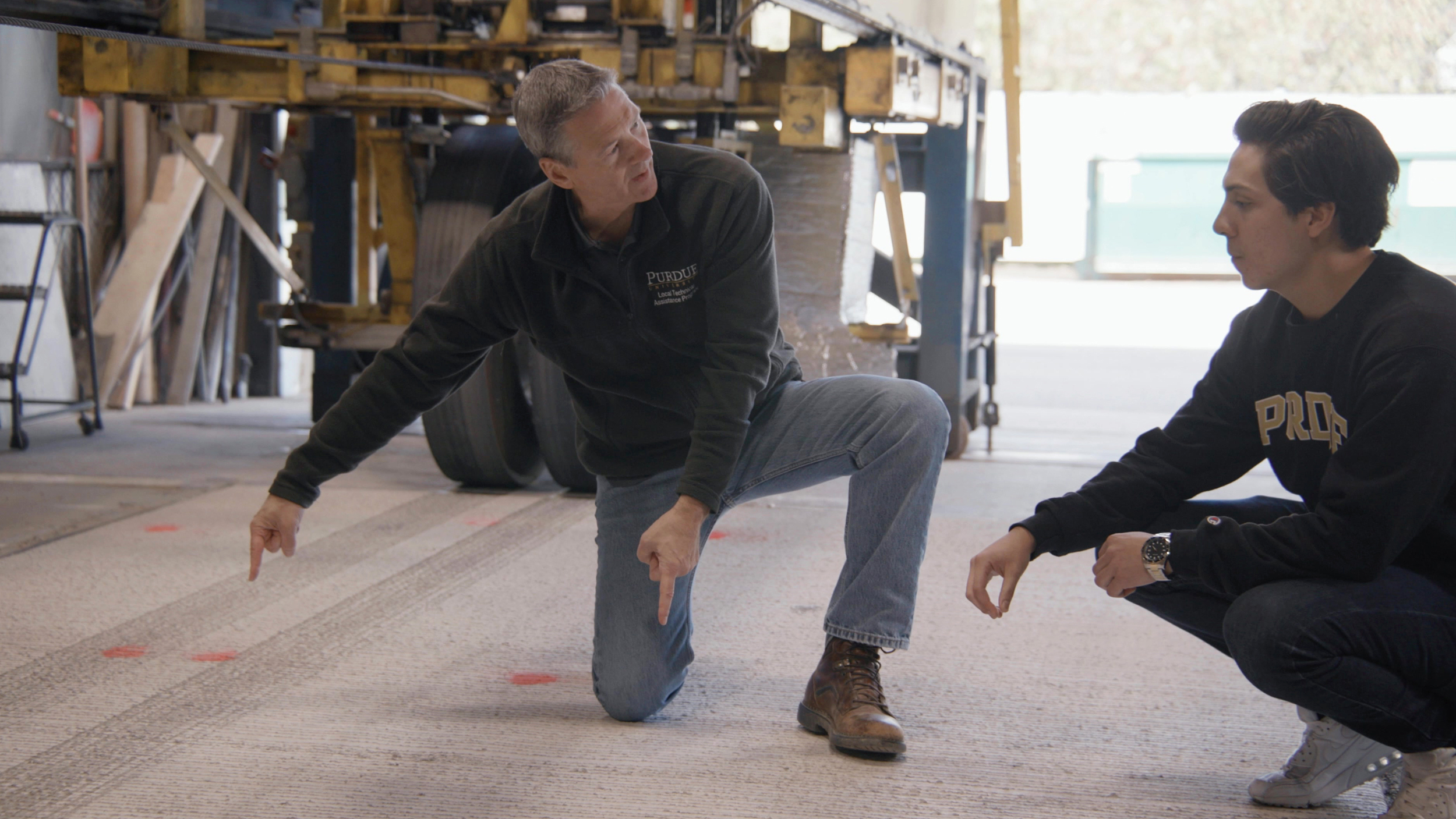A man in jeans, a long sleeved shirt, and boots kneels on the floor and points as another man listens and kneels next to him.
