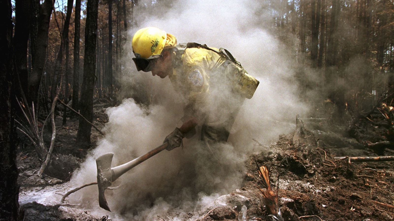 Man in yellow jacket and yellow hard hat hits earth wtih ax during fire