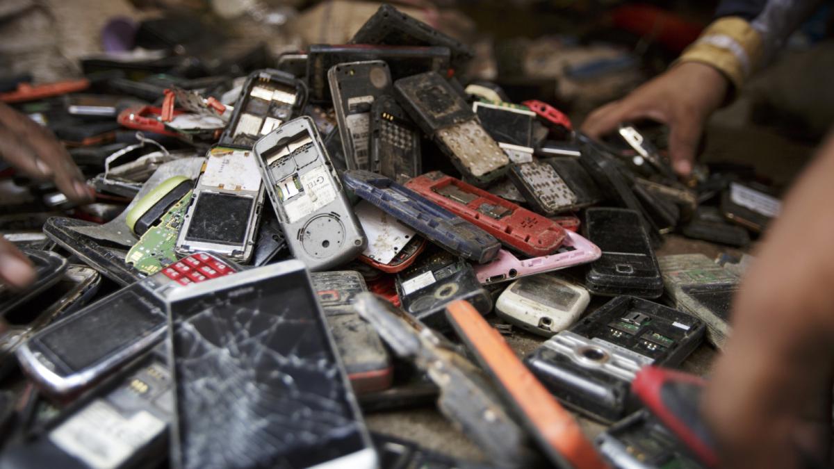 A hand reaches into a large pile of cell phones and other small electronic devices, many of them cracked or missing batteries