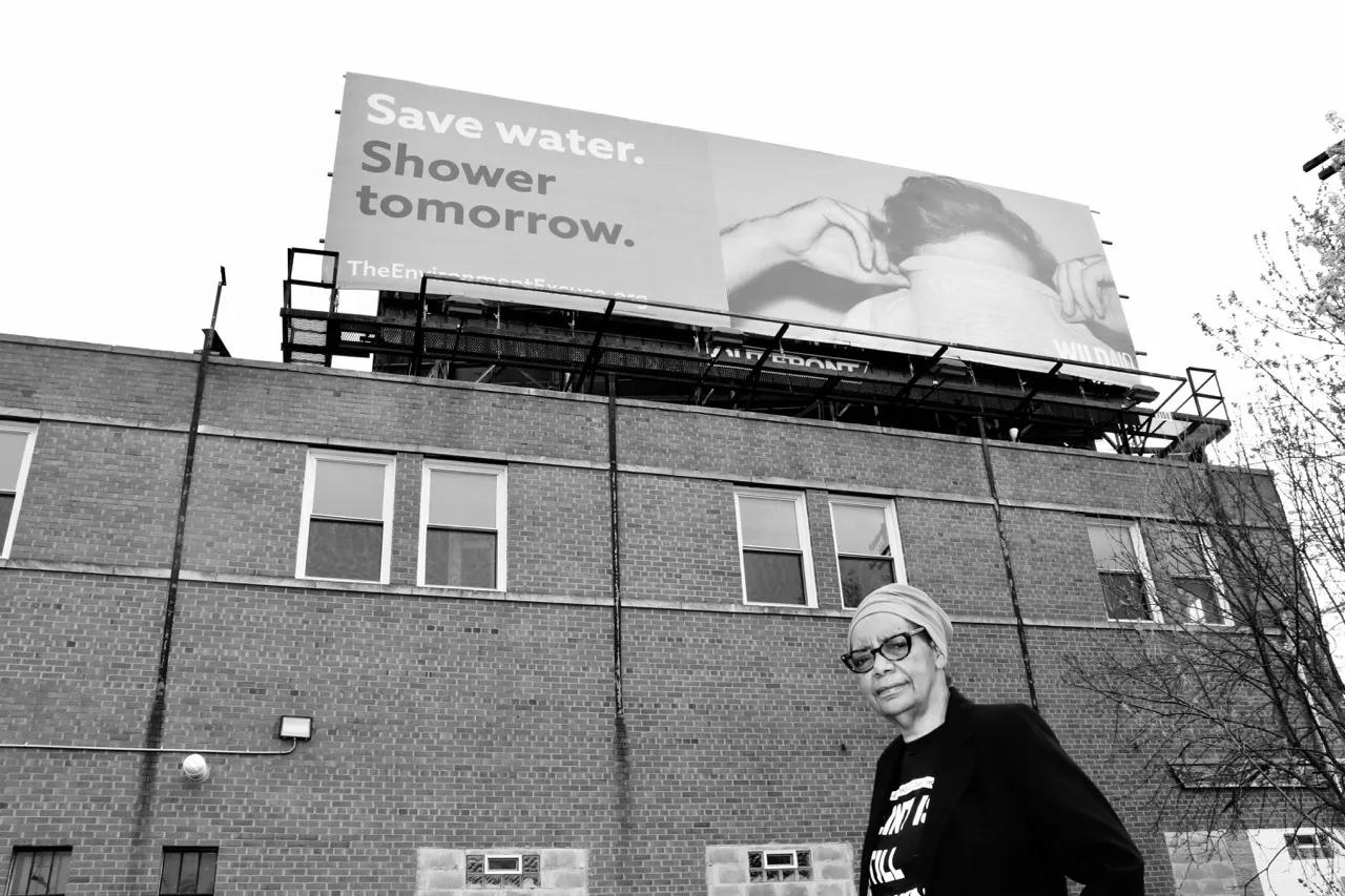 An older woman with glasses and a head wrap walks in front of a brick building with. a billboard that reads Save water. Shower tomorrow.