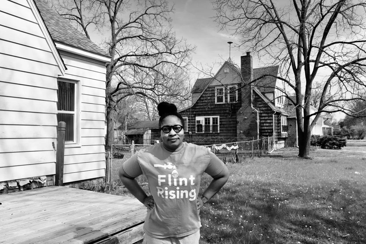 一名黑人妇女身穿印有“弗林特崛起”字样的 T 恤，戴着眼镜，双手叉腰站在自家后院。