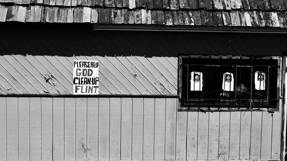A sign that reads Please help God clean up Flint tacked to the side of a building with bars on the windows.
