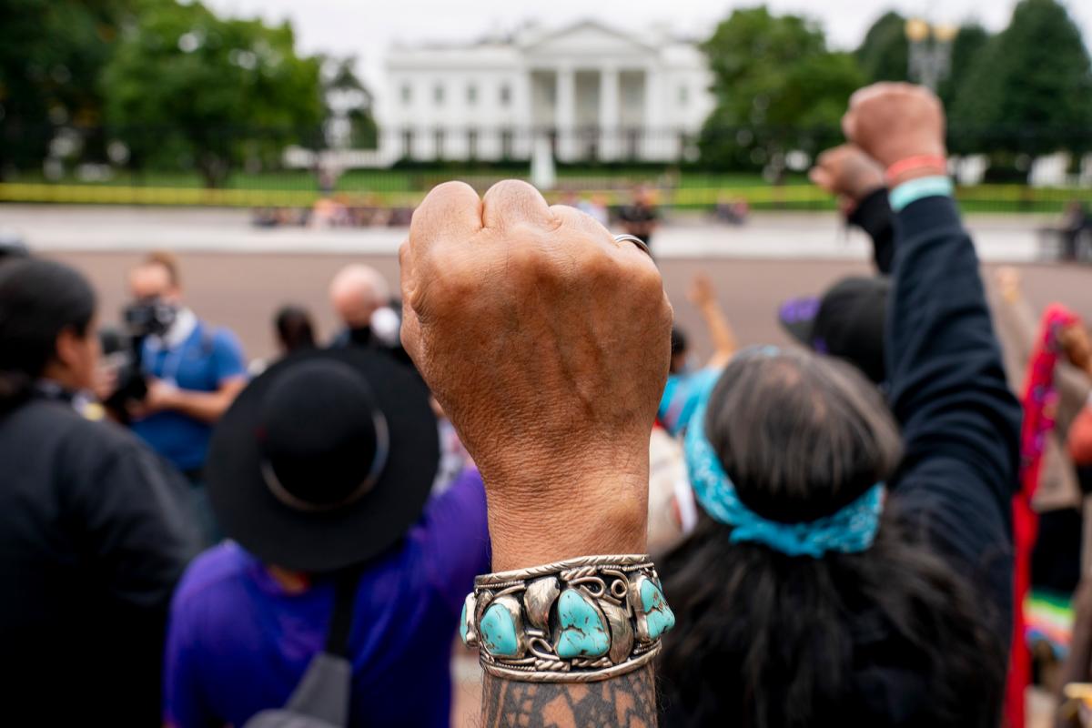 A raised fist of a person wearing a blue and silver bracelet in front of a crowd in front of the white house