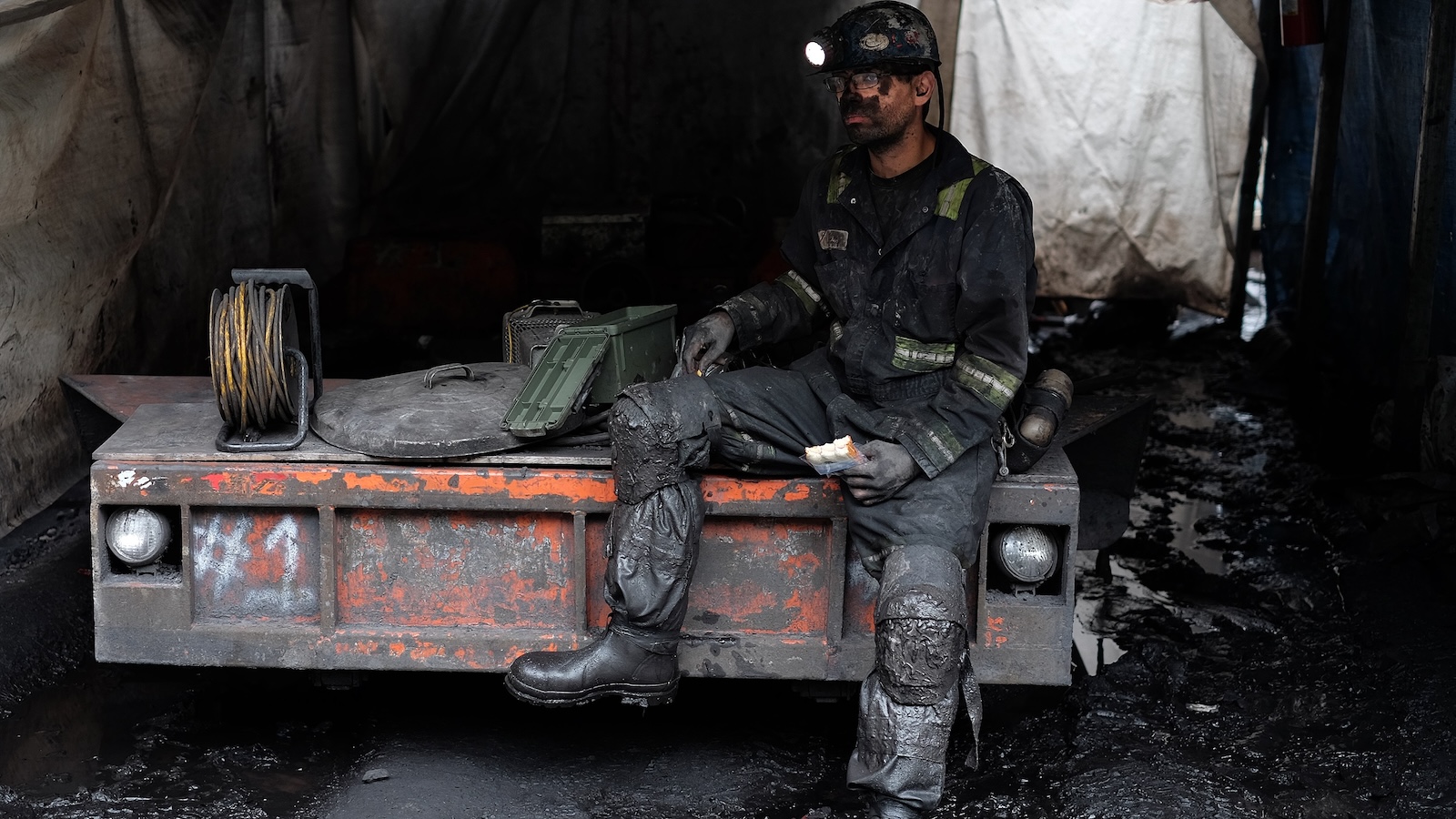 A coal miner wearing coveralls sits on a piece of mining equipment while eating his lunch.