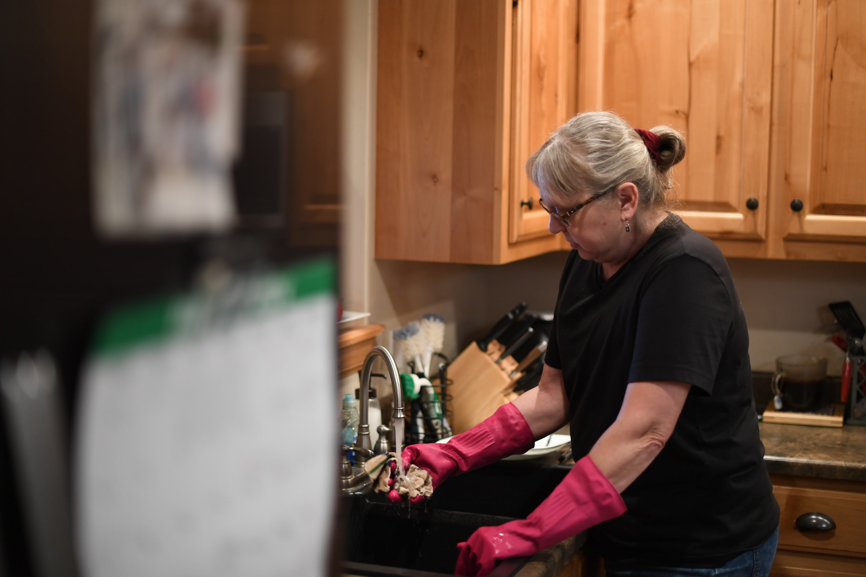 A woman in a black tee shirt with her hair tied back wears red kitchen gloves and stands with her hands in the kitchen sink.