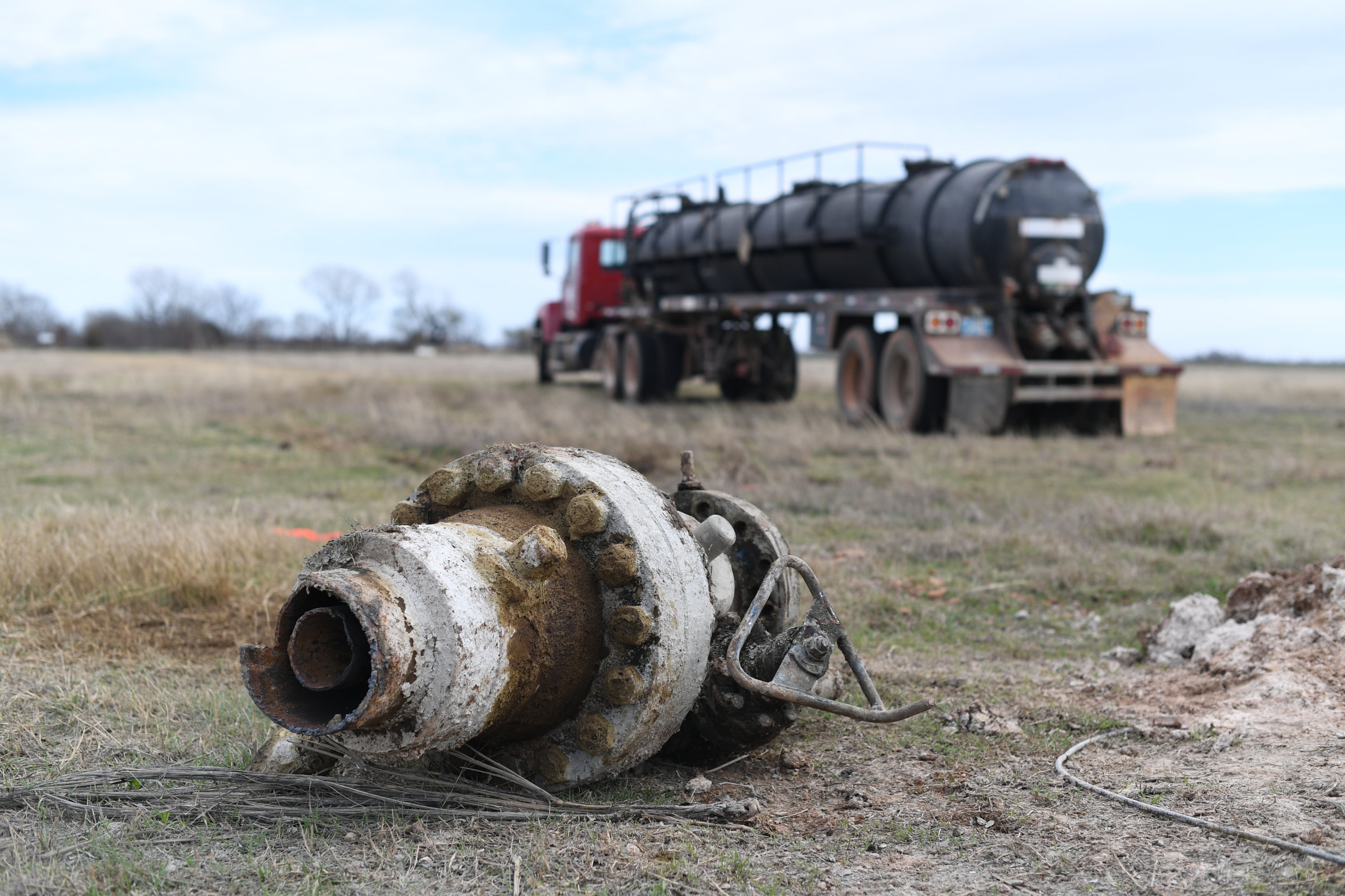 A rusting piece of equipment sits in the gras with a large truck in the background.