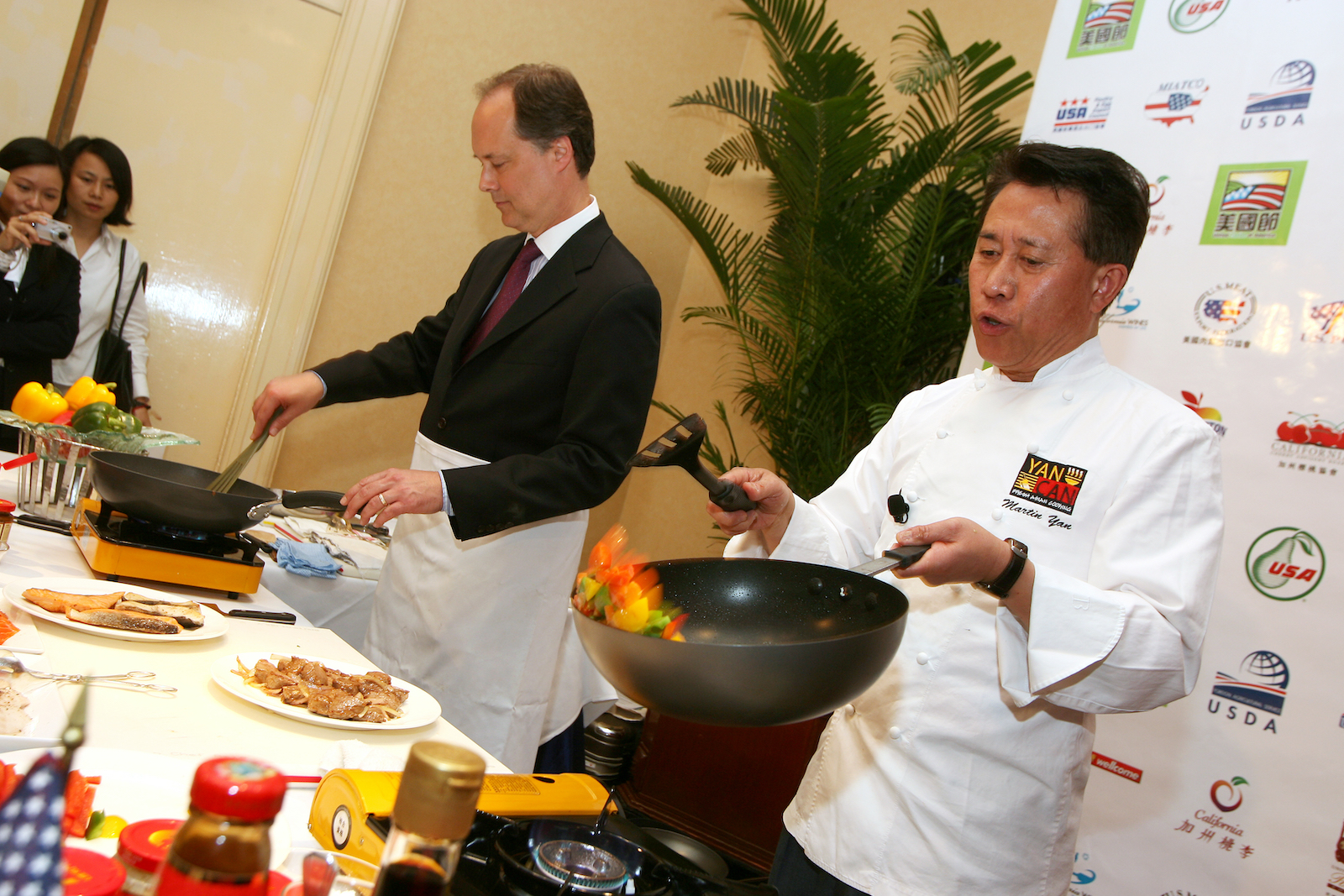 Two men cook over hot stoves. The man on the right wears a white chef uniform and holds a wok with flame
