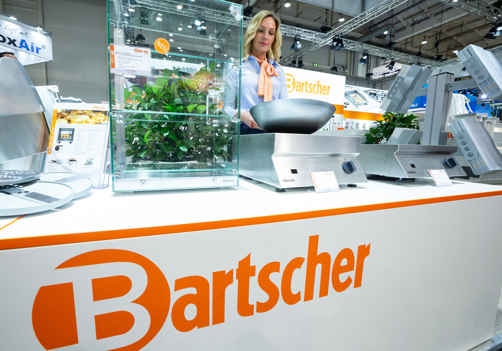 a woman stands in front of a counter labeled Bartscher holding a metal wok on a metal stand induction stove