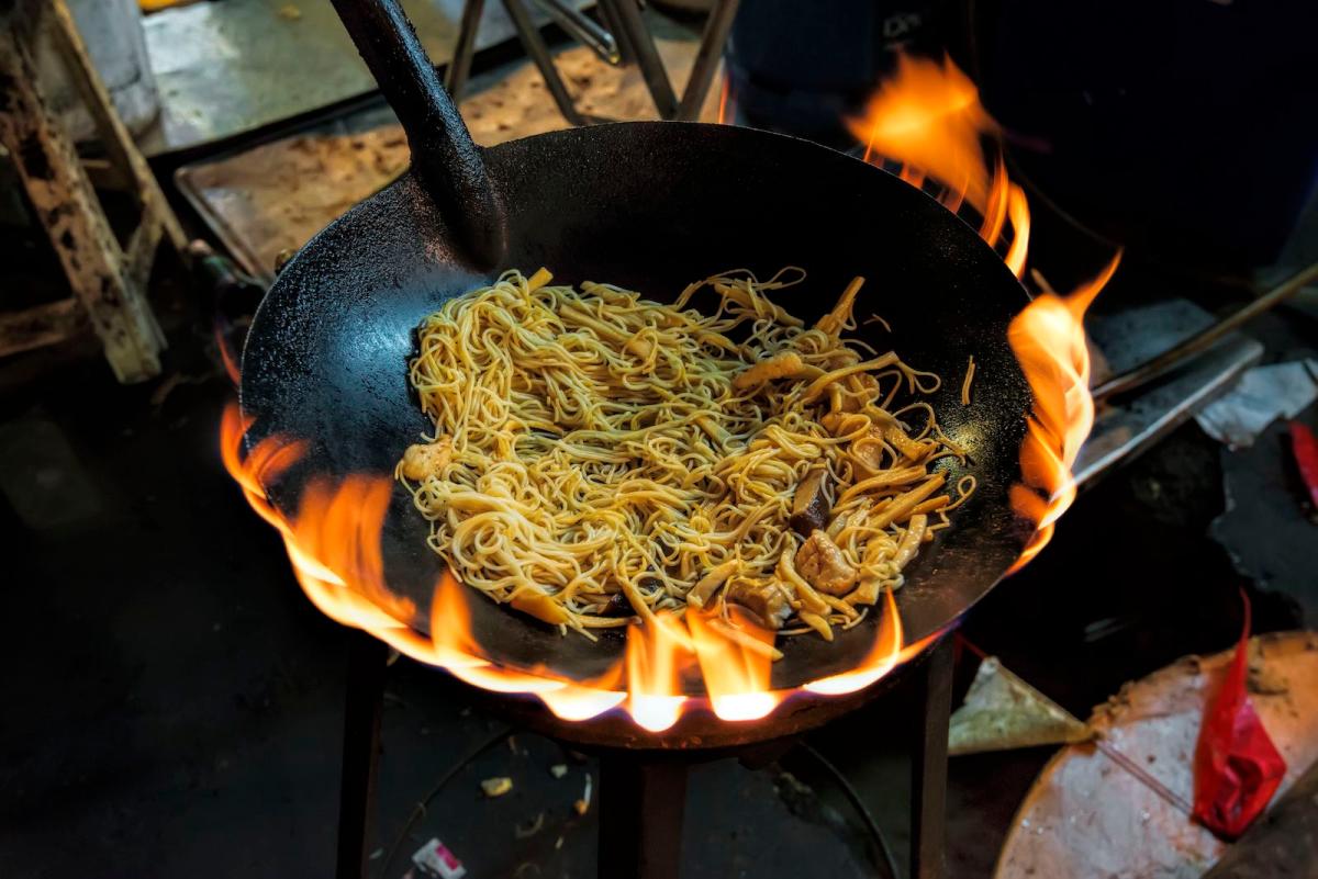noodles cook in a wok over orange flame