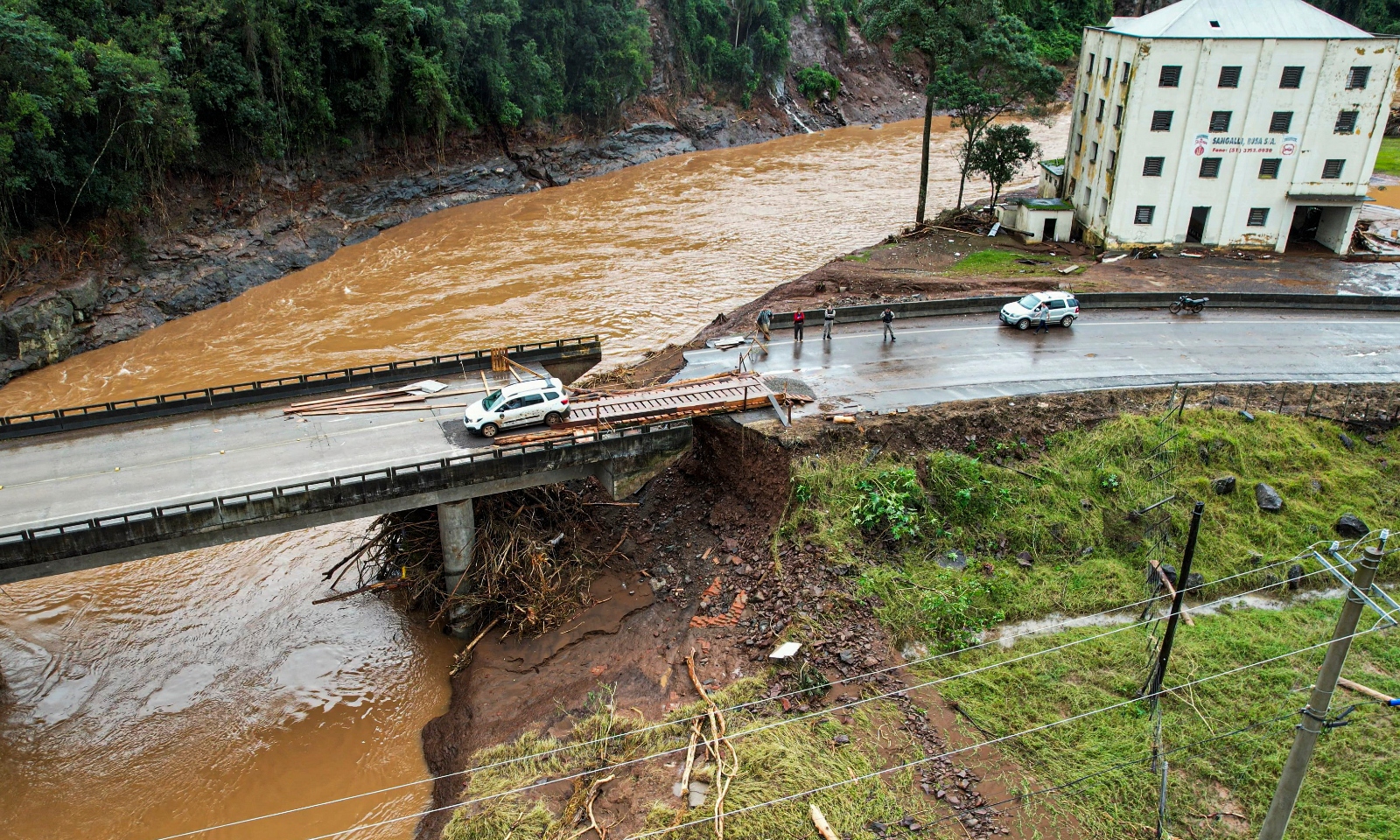 An aerial view of a bridge that crosses over a large brown river. The bridge has been partially destroyed by flooding.