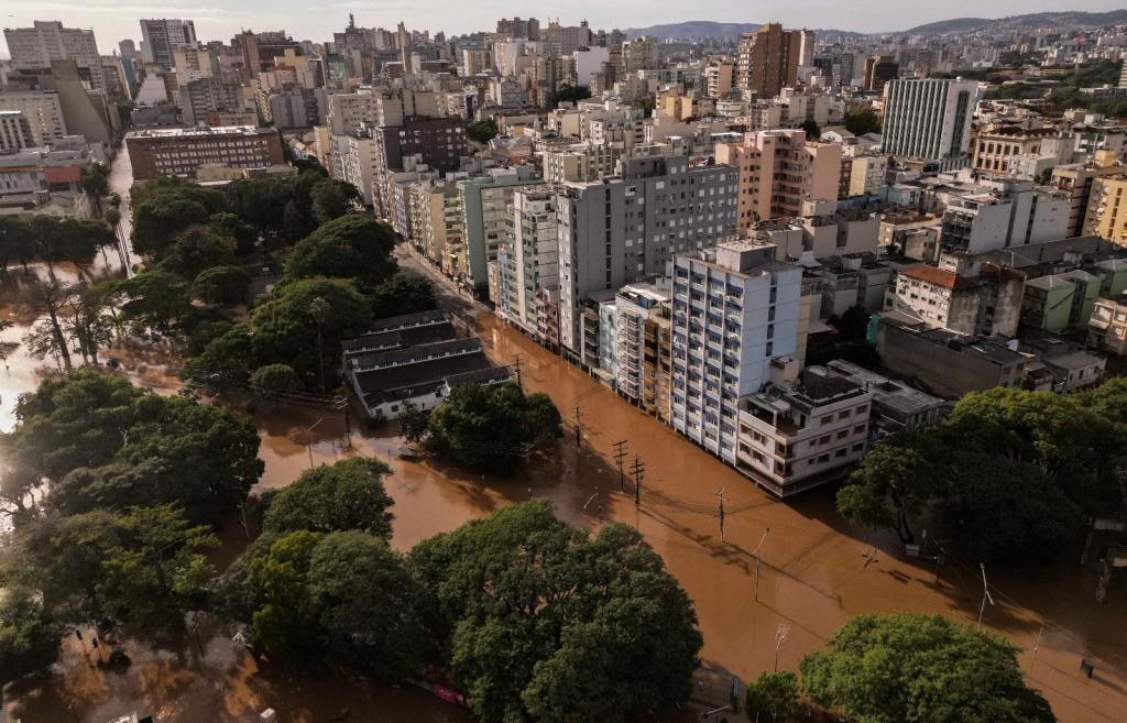 This is an aerial shot of a flooded city neighborhood. The street is lined with white high-rise apartment buildings and the road is submerged in brown floodwater.