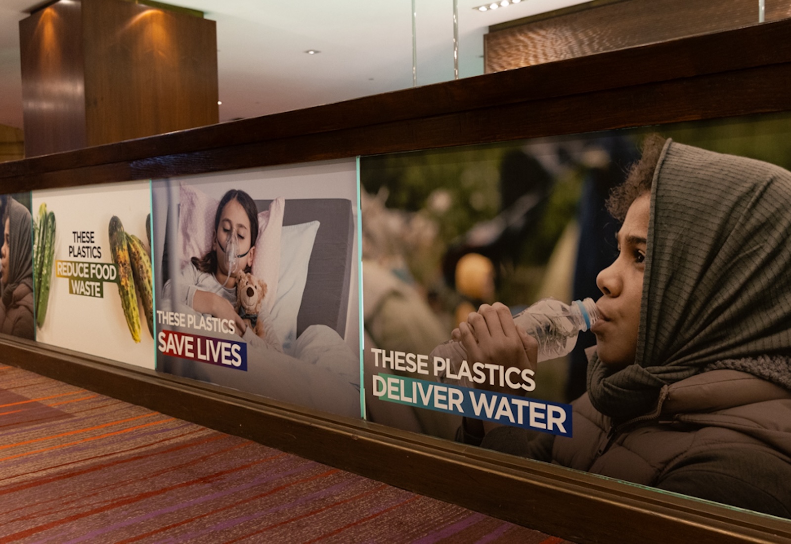 Large ads plastered on walls say 'these plastics reduce food waste' against a photograph of cucumbers, 'these plastics save lives' against a photograph of a child in a hospital bed, and 'these plastics deliver water' against a photograph of a child drinking from a disposable water bottle