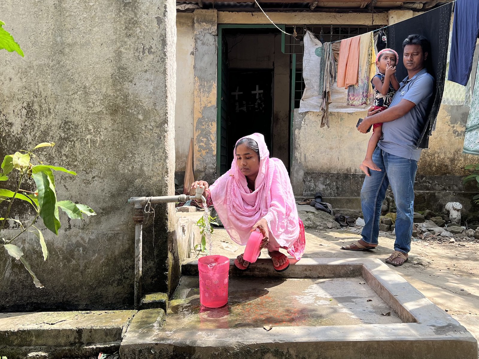 Wearing pink from head to toe, Akhter fills pink pitchers with water from a spigot in front of her house. Her husband, Shamim, stands nearby holding their daughter, Muntaha, in his arms.