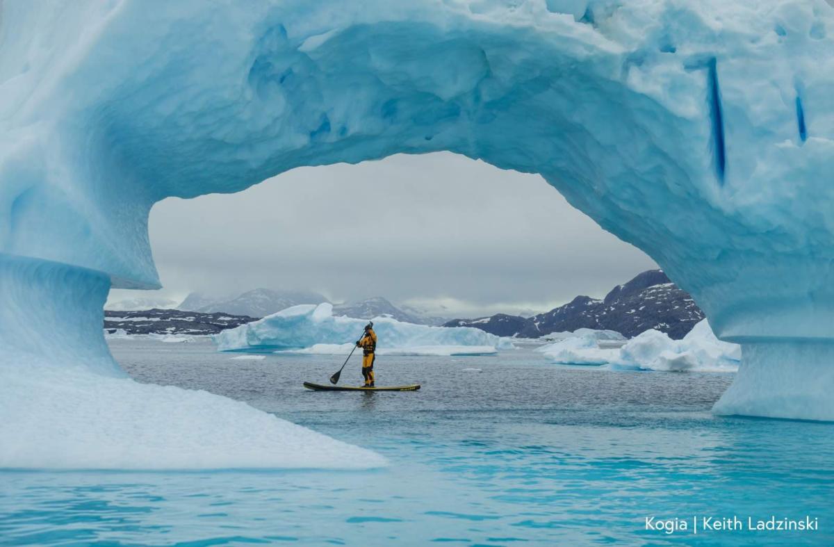 A view through an ice arch shows a person on a stand-up padleboard, surrounded by ice floes