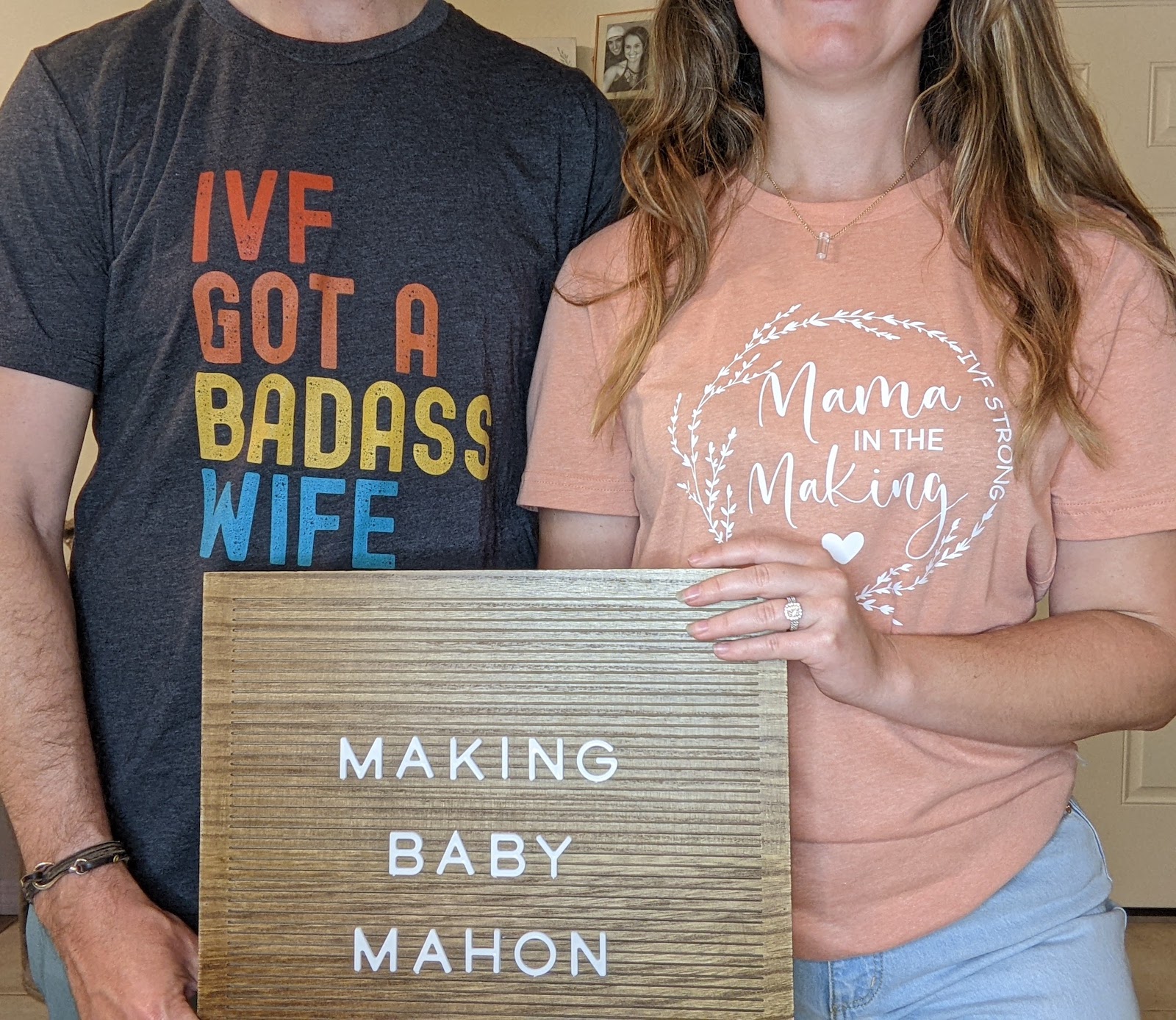 A couple wear shirts that say 'ivf got a badass wife' and 'mama in the making' in front of a wooden sign that says 'making baby mahon'