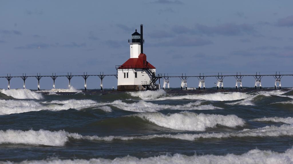 red-roofed lighthouse stands in rolling blue water with white waves against blue-gray sky
