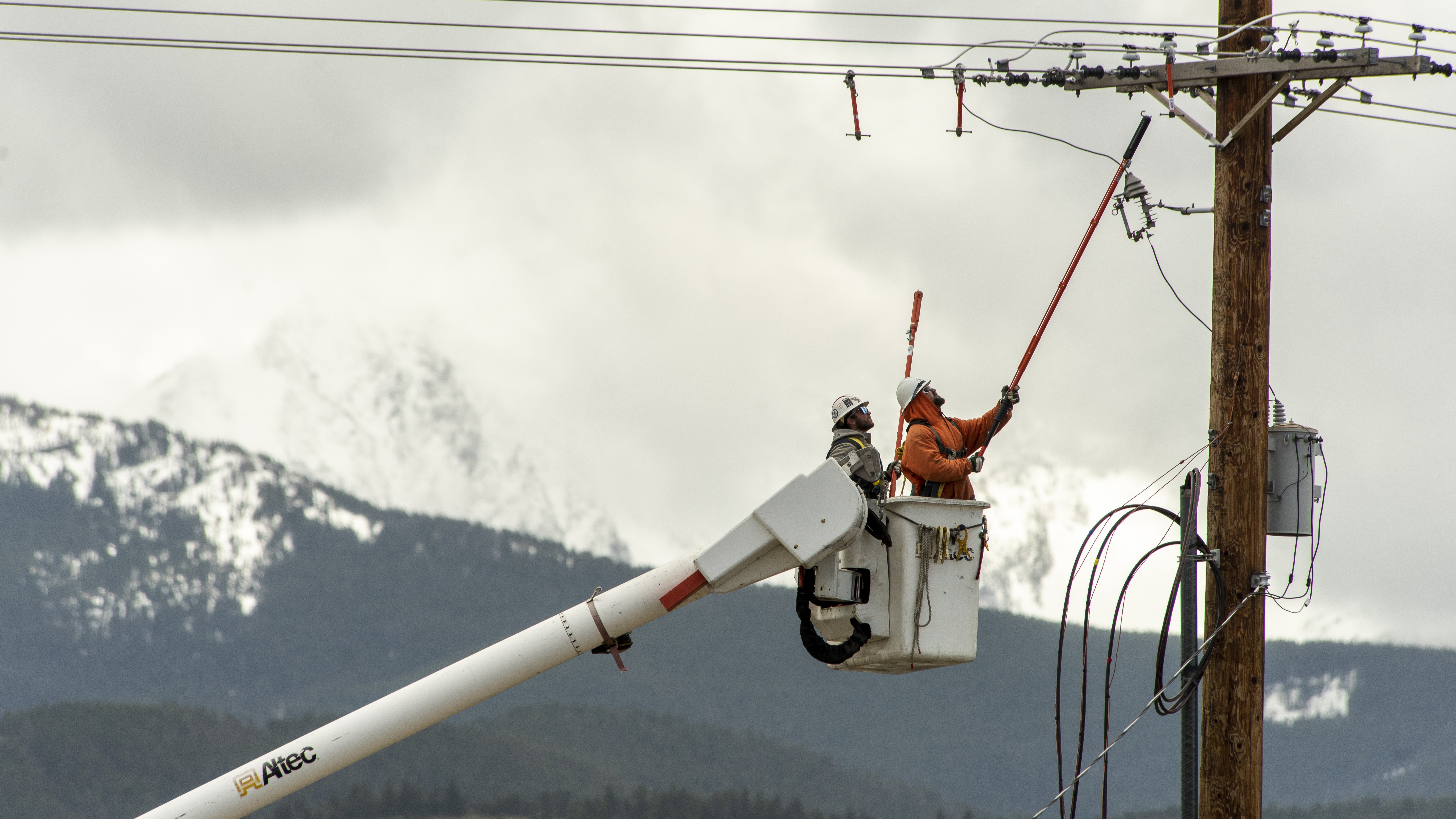 Electric workers in a boom lift work on a transmission line with a snow-capped mountain in the background.