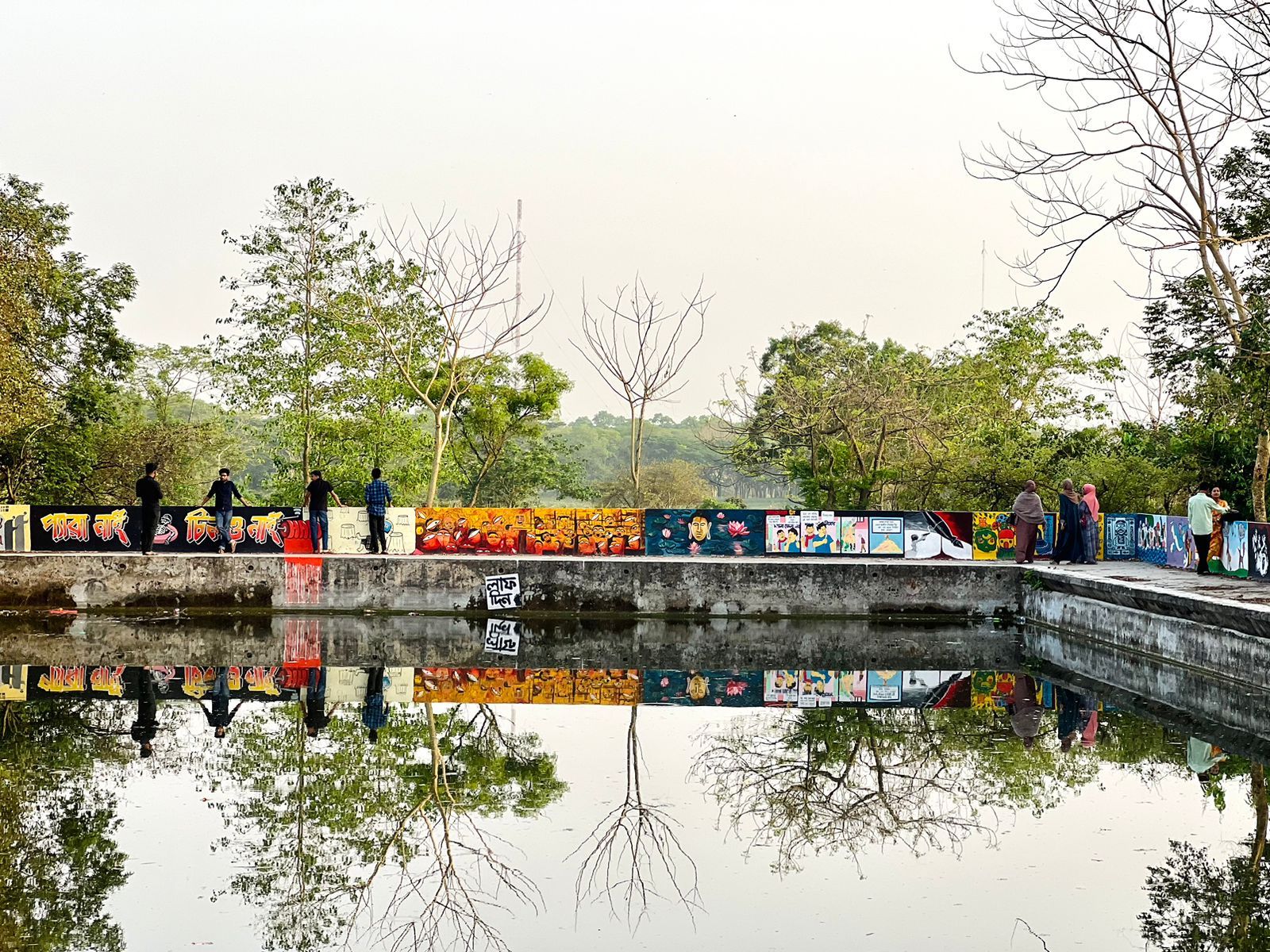 People stand near a reflective reservoir surrounded by trees in an urban setting