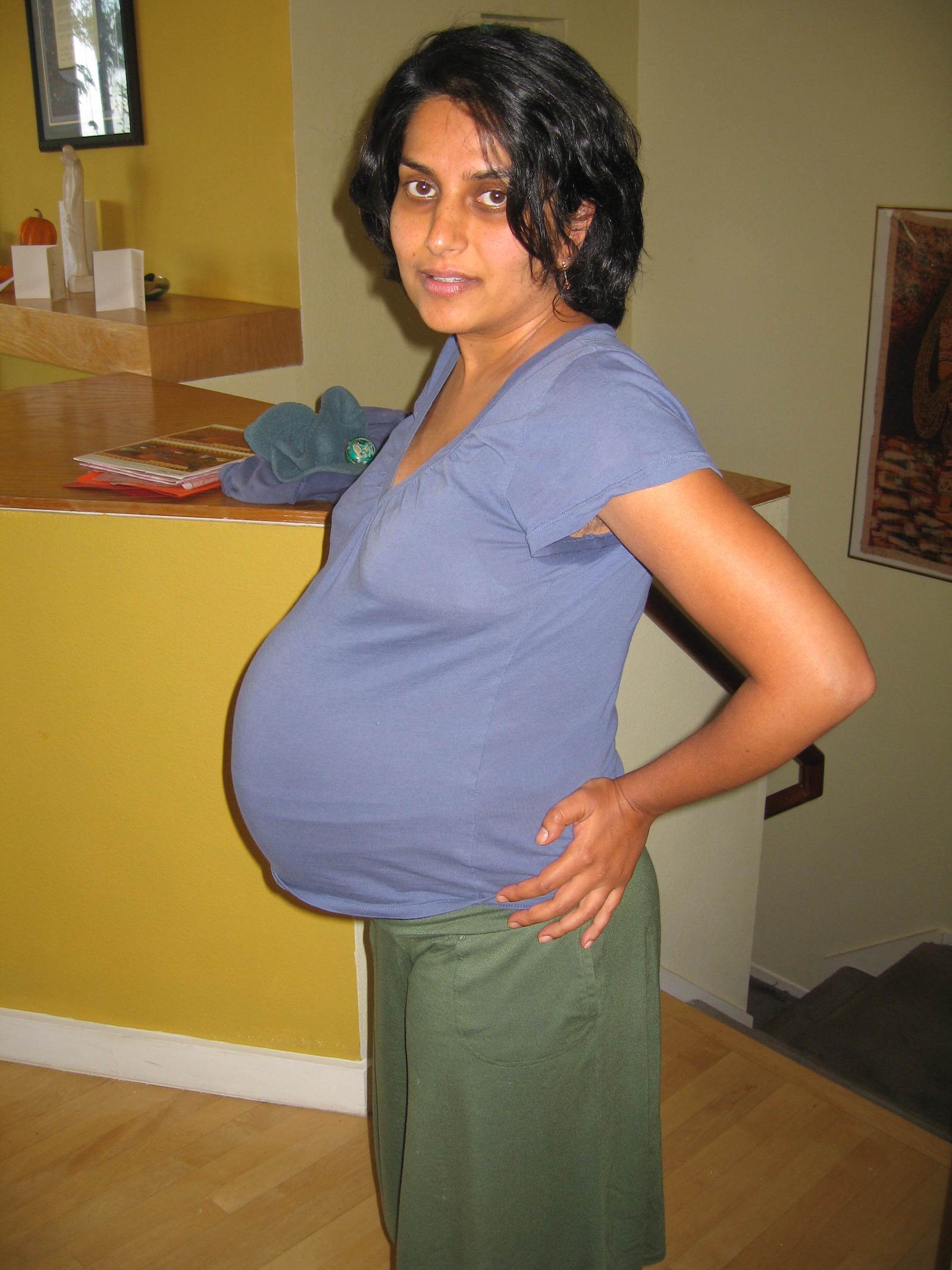 A woman in a blue-t-shirt and with a large pregnant belly puts her hands on her hips and looks into the camera