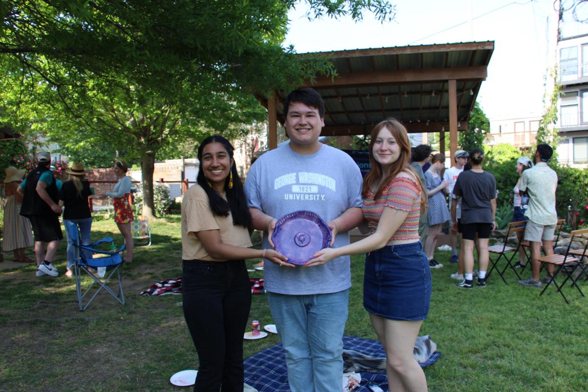 Three smiling people stand in a grassy area holding up a purple ceramic birdbath