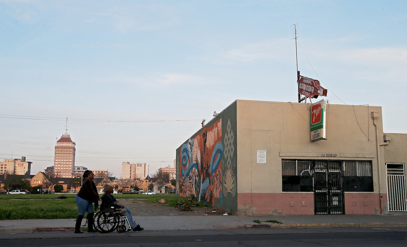 A woman pushes a person in a wheelchair down a city street near a mural
