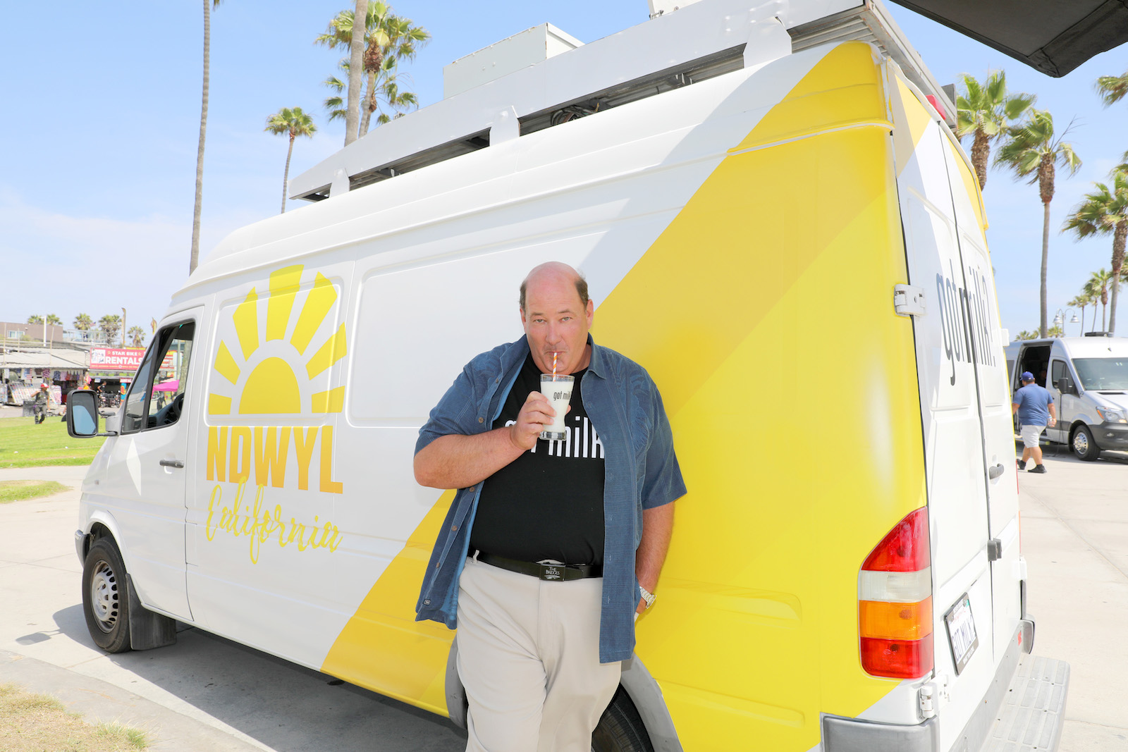 A balding man in a got milk shirt stands in front of a yellow van while drinking milk