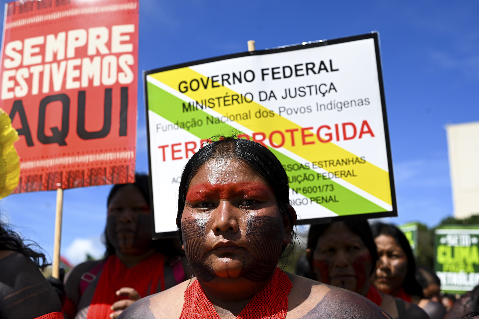 A woman with intricate tattoos and red face paint stares directly into the camera with protest signs behind her