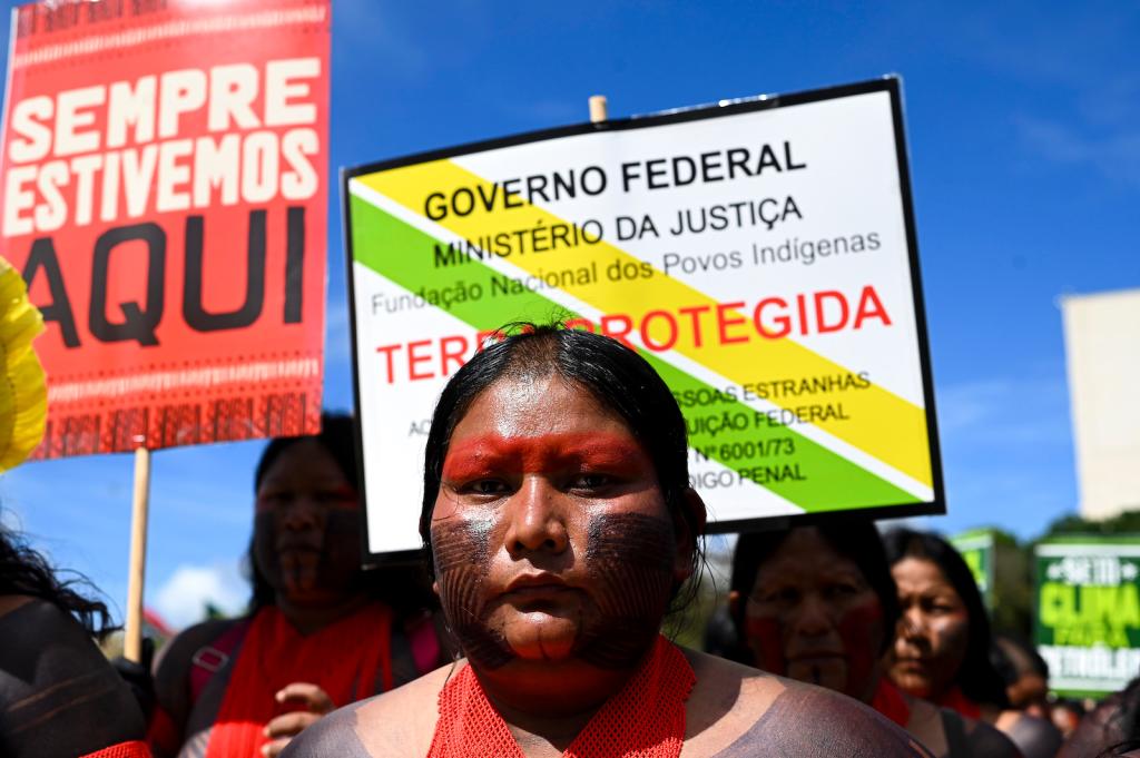 A woman with intricate tattoos and red face paint stares directly into the camera with protest signs behind her