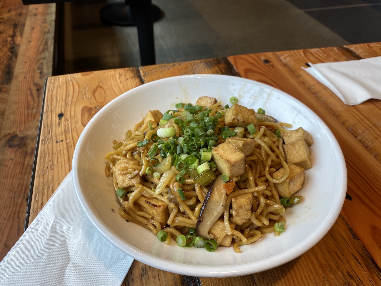A plate of stir fry noodles with tofu sitting on a wooden table with napkins