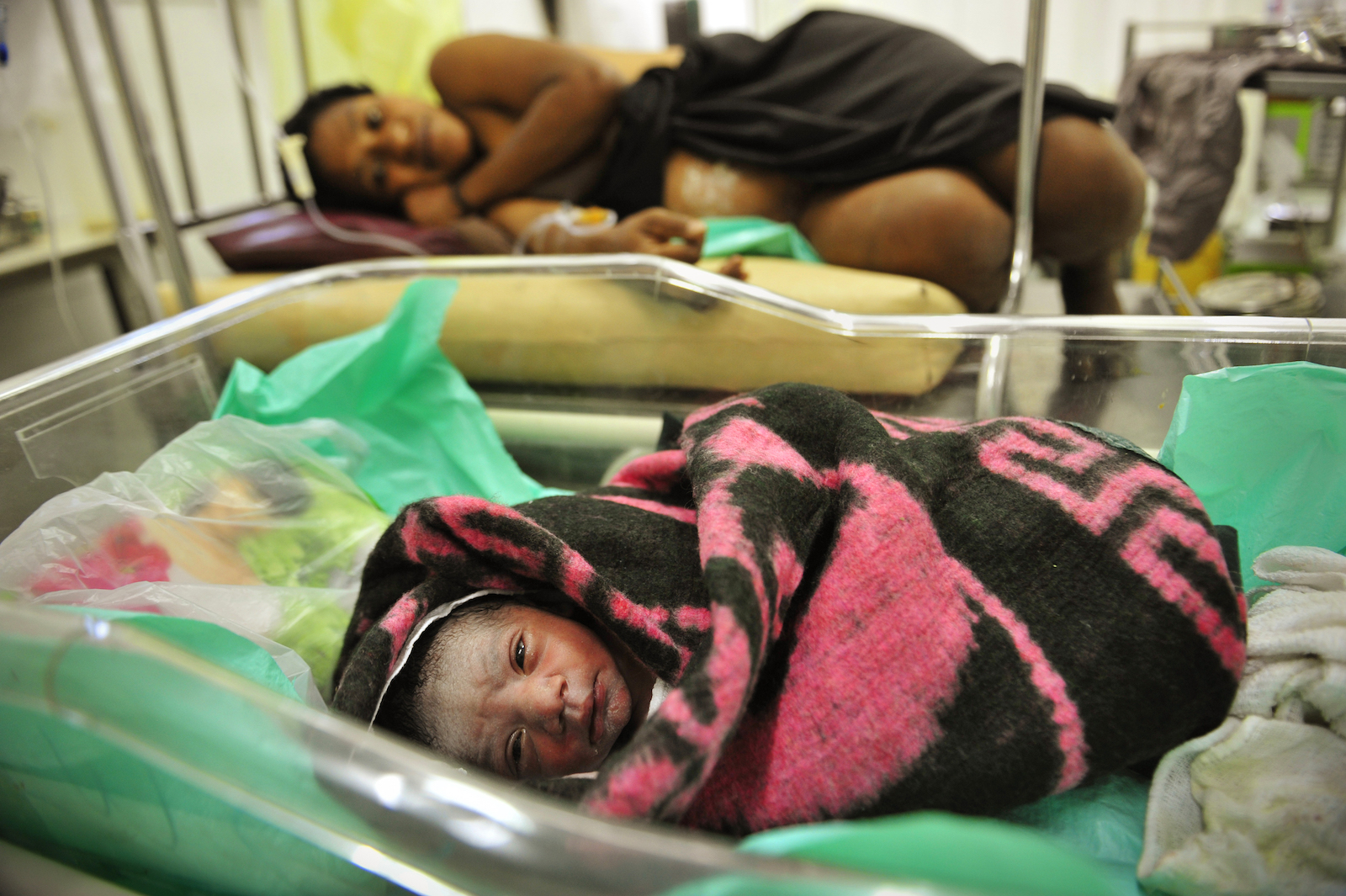 A woman in a hospital bed lays next to a swaddled newborn