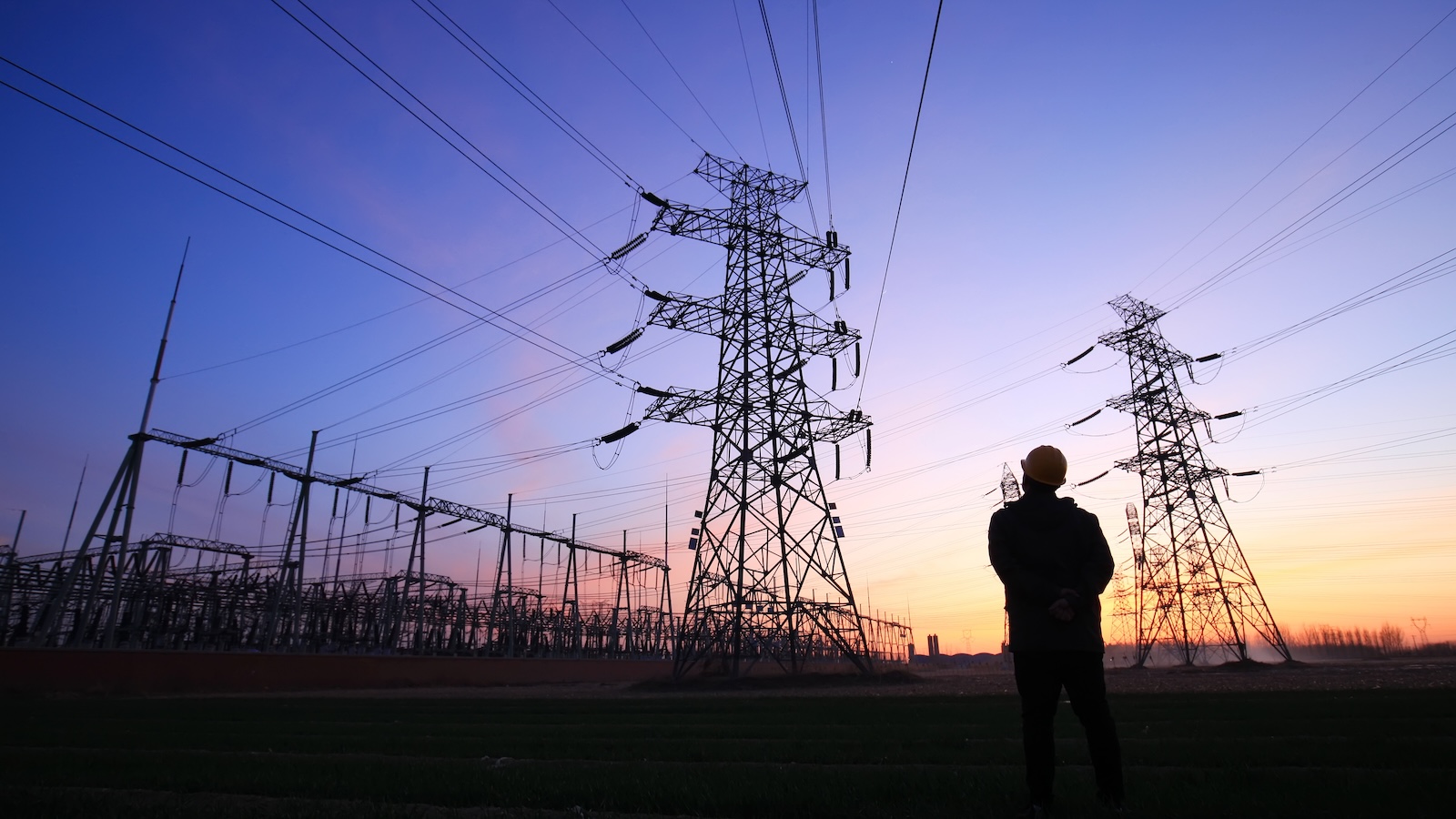 An electric utility worker inspects a transmission tower at dusk.
