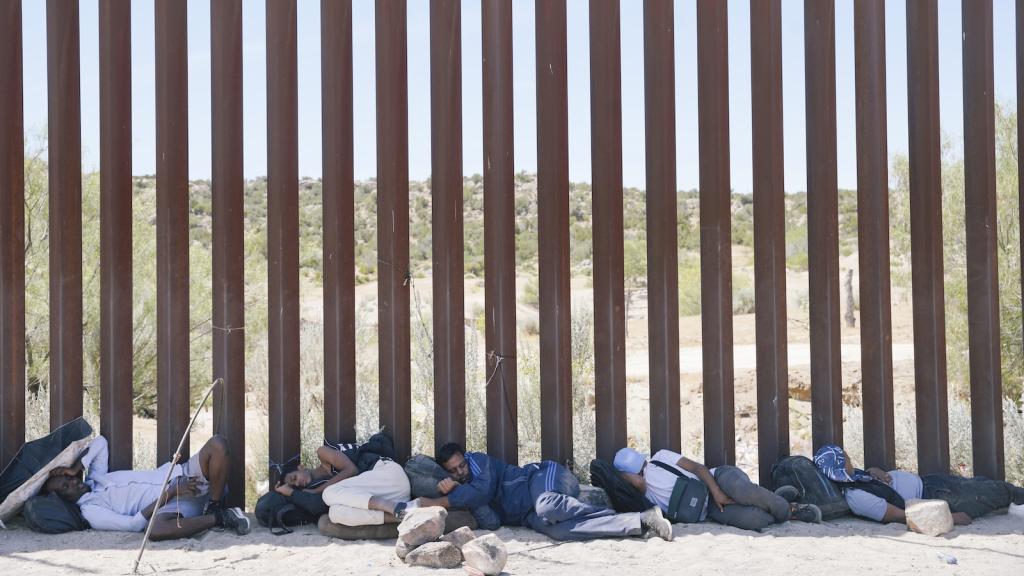 A line of migrants sleeps in a sliver of shade provided by the border wall separating the US and Mexico.