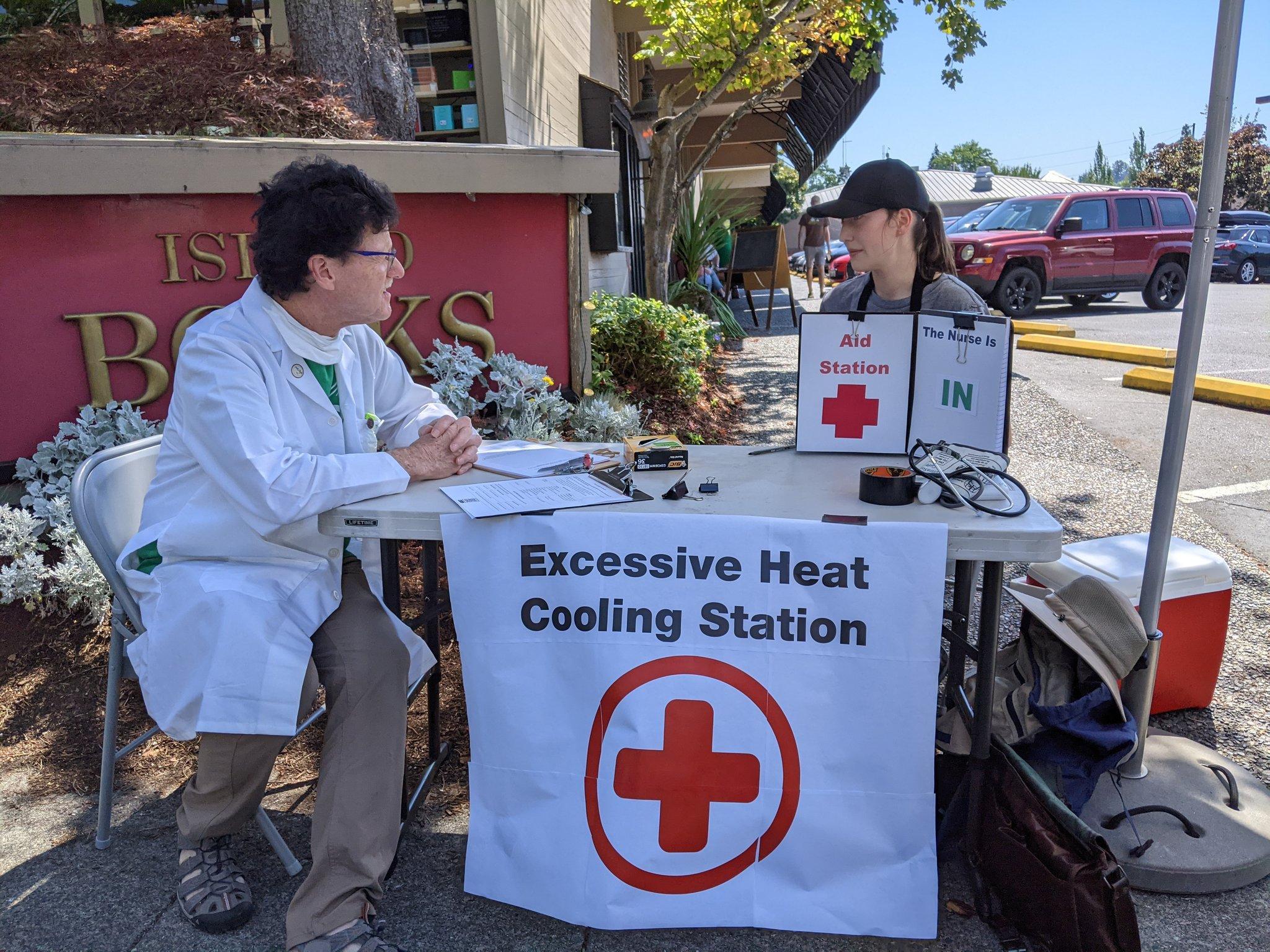 A man and a woman sit down at a table with banner hanging on the front that says 'Excessive Heat Cooling Station'