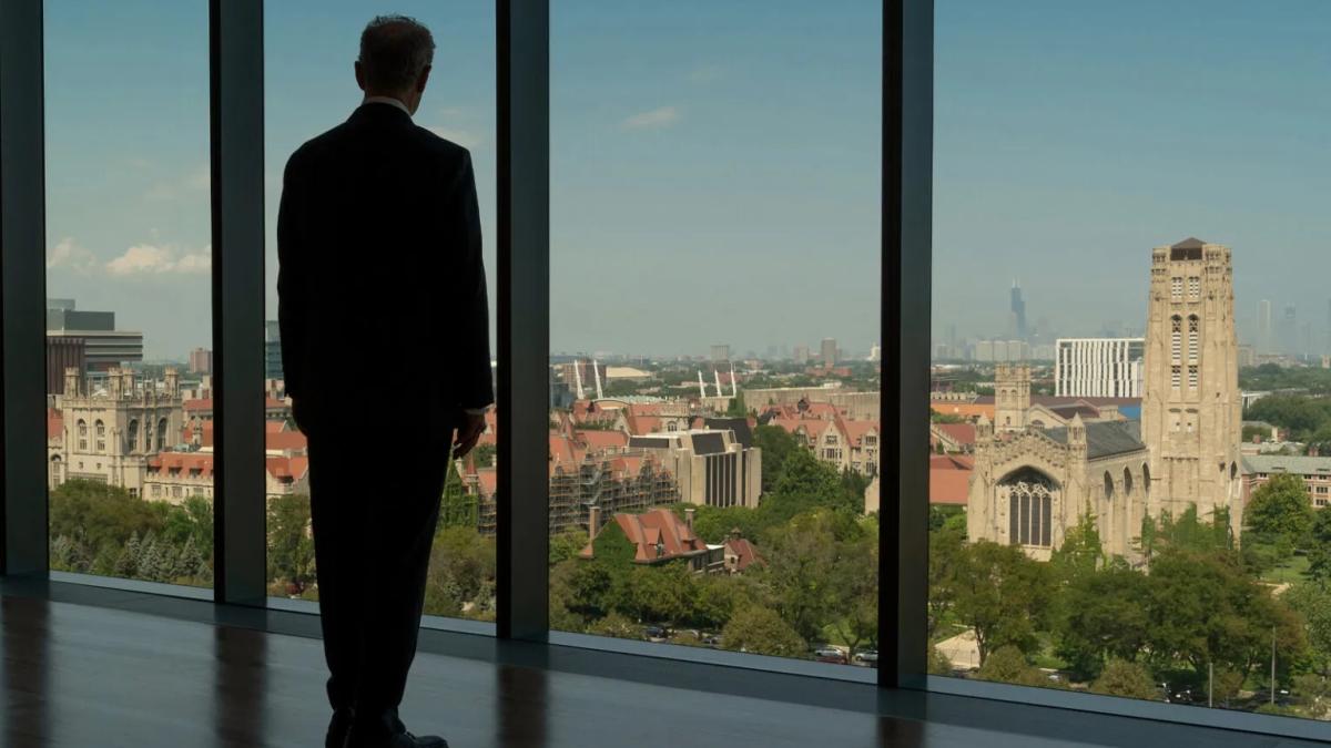 A man in a suit looks out at floor to ceiling windows over a college campus.