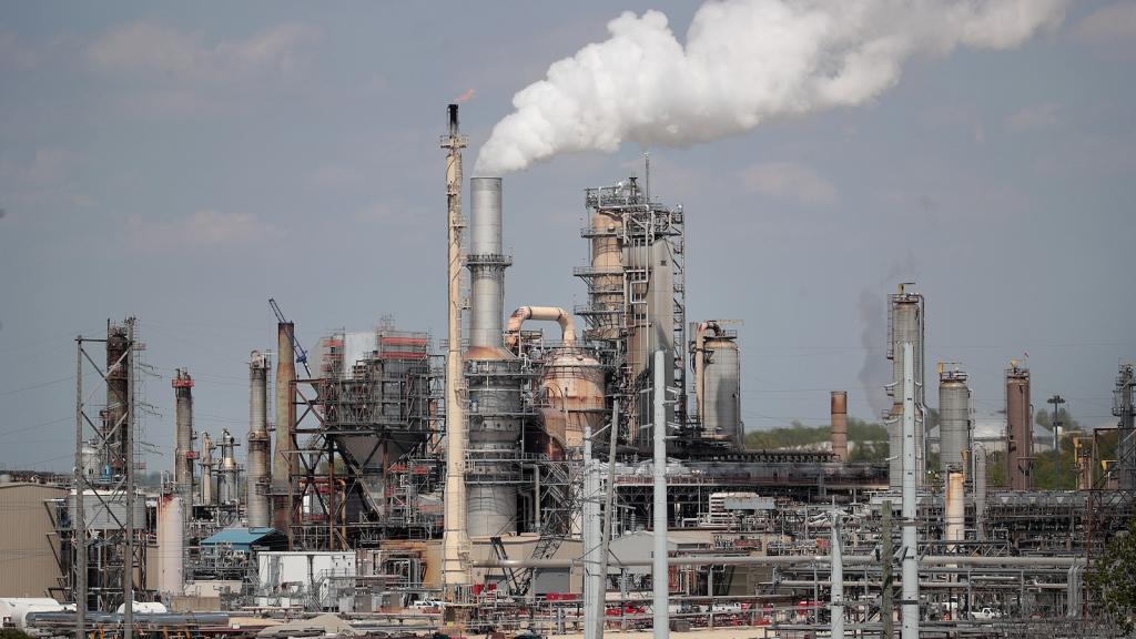 An array pf tall steel smokestacks and pipes with one central smokestack that is billowing white smoke.