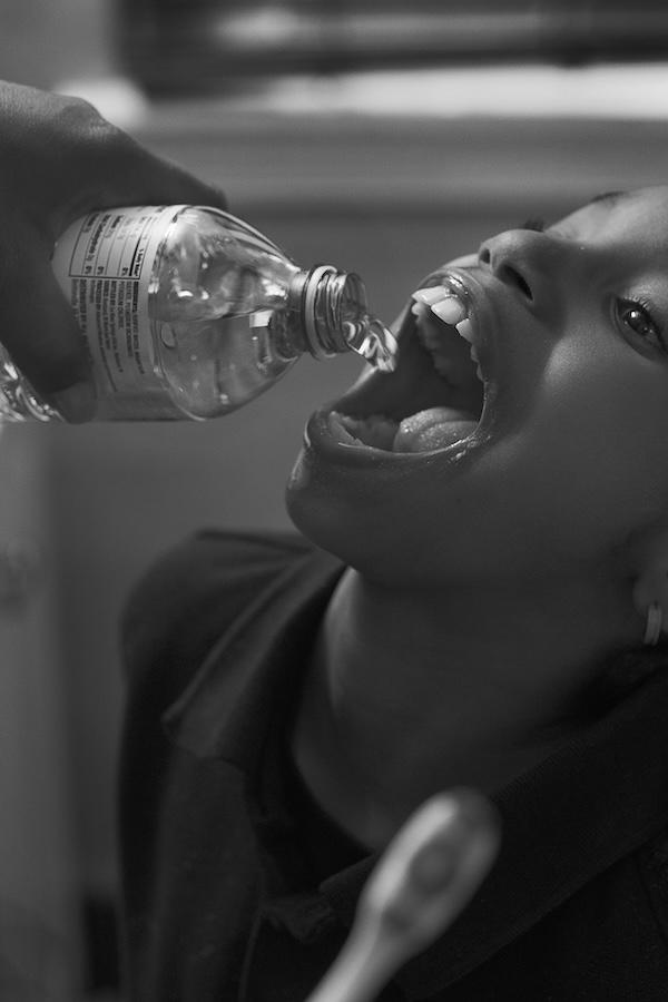 A black and white photo of a child drinking bottled water poured by another person into her mouth