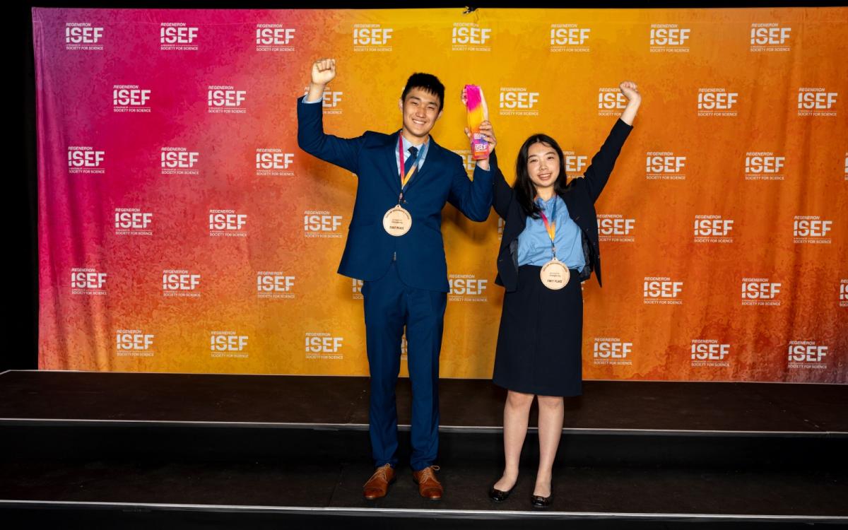 Two smiling teenagers hold up their hands with medals around their necks, standing in front of a curtain with ISEF written on it