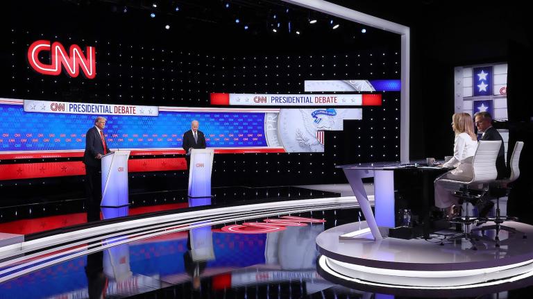 President Joe Biden and former president Donald Trump stand behind podiums on the left, and behind them is a large blue screen with the words "Presidential Debate." Moderators sit at a desk on the right.