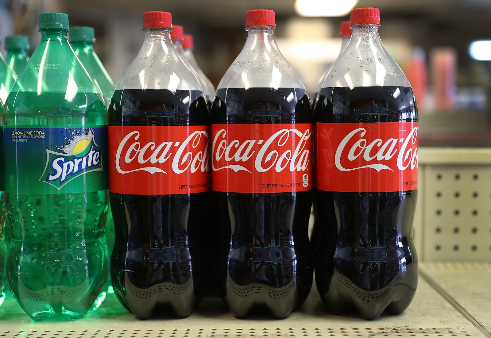 Bottles of Coca-Cola are lined up on a shelf. To the left are large bottles of Sprite.