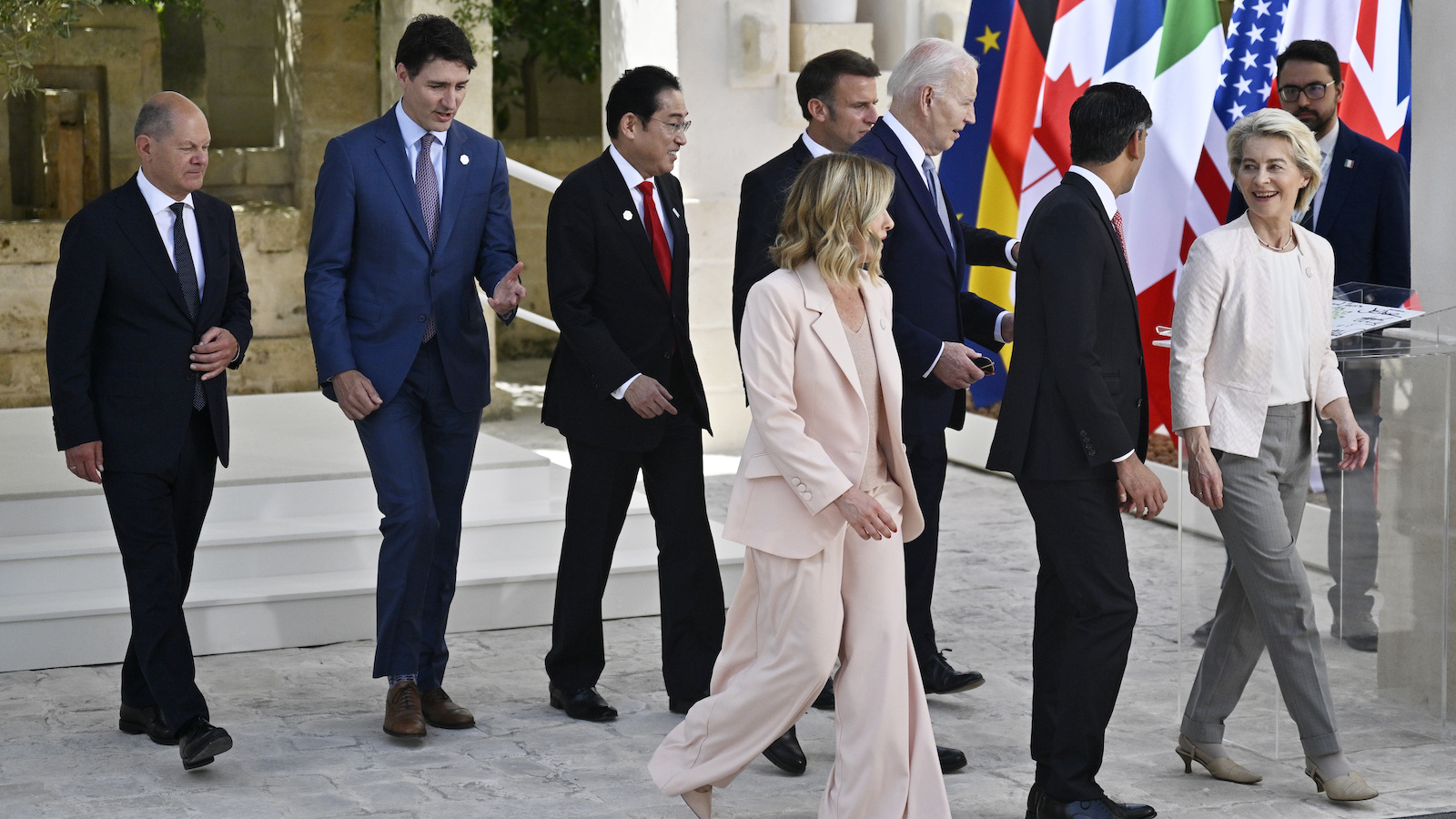 Seven men in suits and two women in pantsuits walk in the same direction across some gray cobblestones with seven world flags behind them