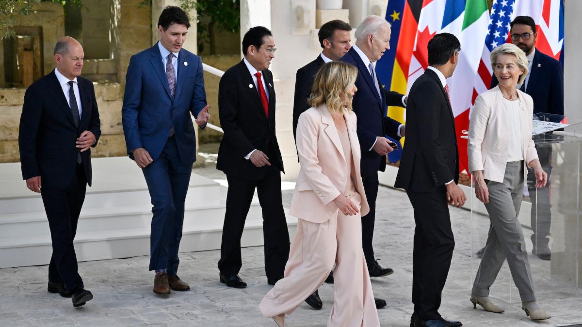 Seven men in suits and two women in pantsuits walk in the same direction across some gray cobblestones with seven world flags behind them