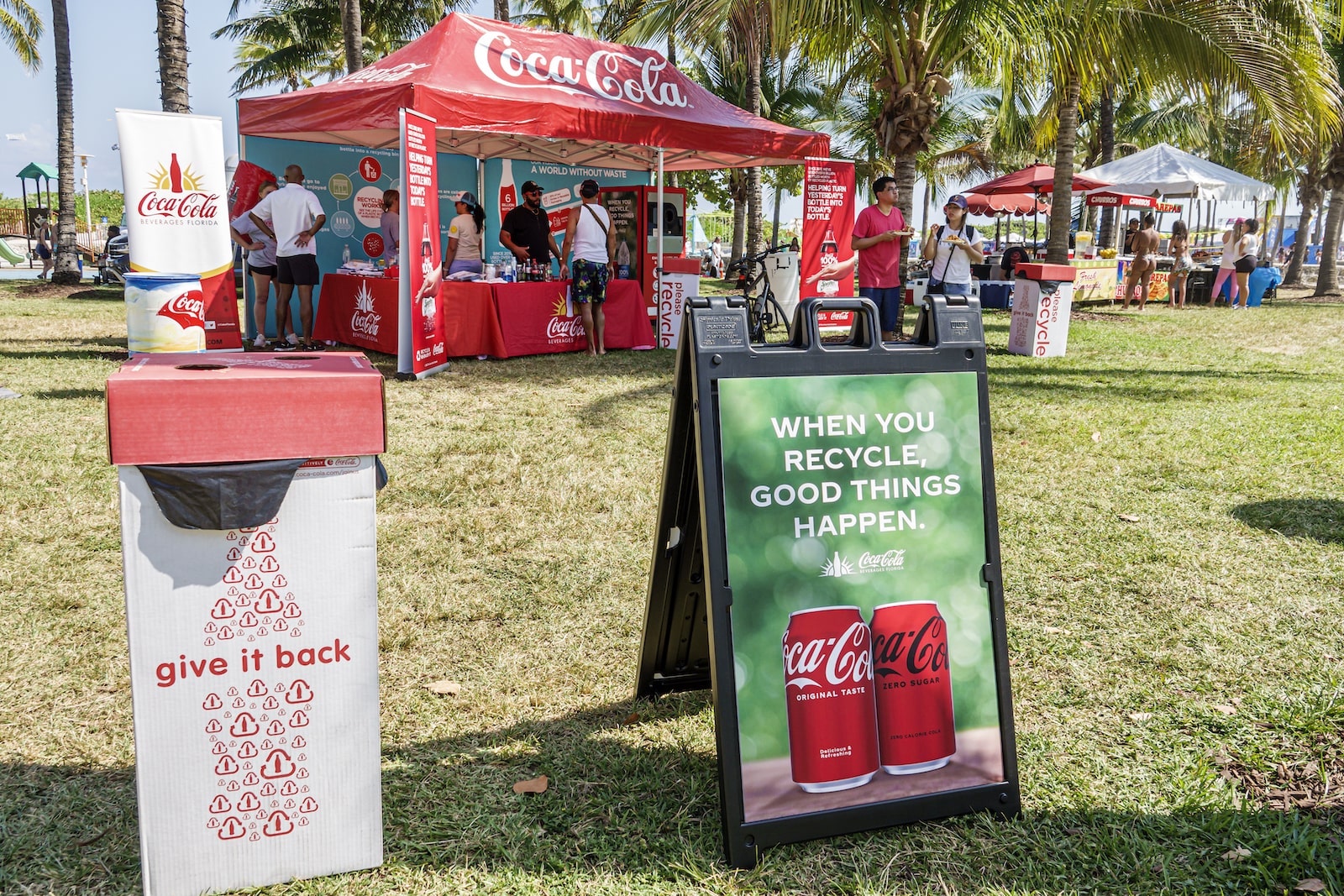 a coca cola kiosk on a green lawn next to a sign that says 'when you recycle, good things happen' with coca cola images