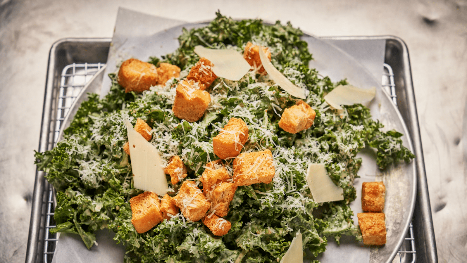 A vegan kale Caesar salad dropped with croutons and shaved and grated vegan cheese sits on a plate on a wire rack.